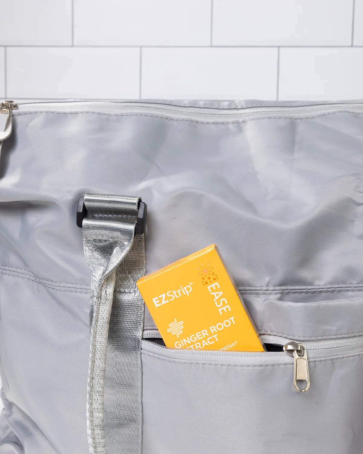 EZStrip Ginger Root Extract dietary supplement package partially placed inside the pocket of a stylish gray travel bag, positioned against white tiled background.