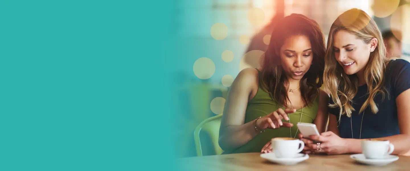 Two young women enjoying each other's company at a cozy café. They are both smiling as they engage with a smartphone, perhaps sharing content or messages, with two cups of coffee placed on the table in front of them. Warm, cheerful lighting enhances the casual and relaxed atmosphere.