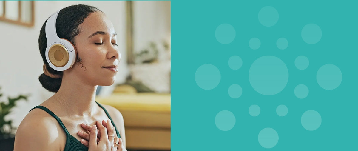Peaceful young woman practicing meditation indoors, wearing white and gold headphones, eyes gently closed, and hands softly placed on her chest. The quiet, calming expression on her face suggests deep relaxation, mindfulness, and tranquility within a comfortable home setting.