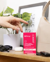 A hand reaching for a bright pink box of EZStrip Boost dietary supplement with Camu Camu and Elderberry Extracts, placed next to a beige purse and sunglasses.