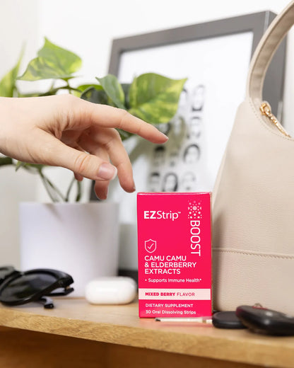 A hand reaching for a bright pink box of EZStrip Boost dietary supplement with Camu Camu and Elderberry Extracts, placed next to a beige purse and sunglasses.