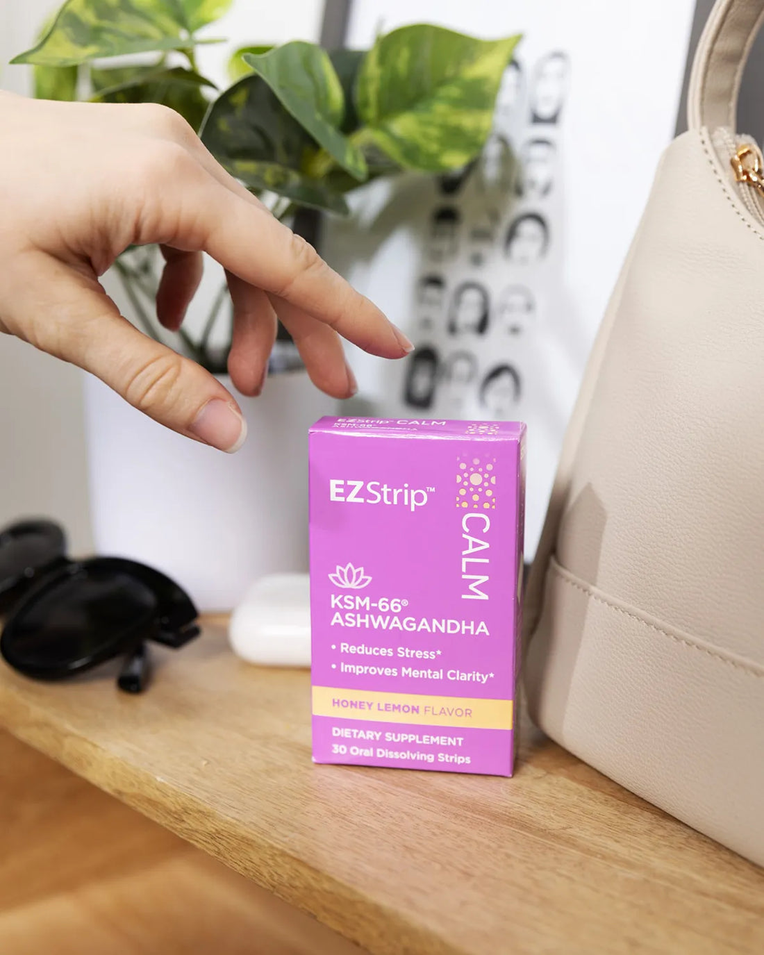 A hand reaching for a purple box of EZStrip Calm dietary supplement with KSM-66 Ashwagandha, placed next to a beige purse and sunglasses.
