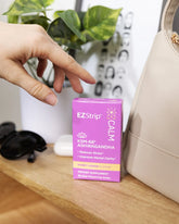 A hand reaching for a purple box of EZStrip Calm dietary supplement with KSM-66 Ashwagandha, placed next to a beige purse and sunglasses.