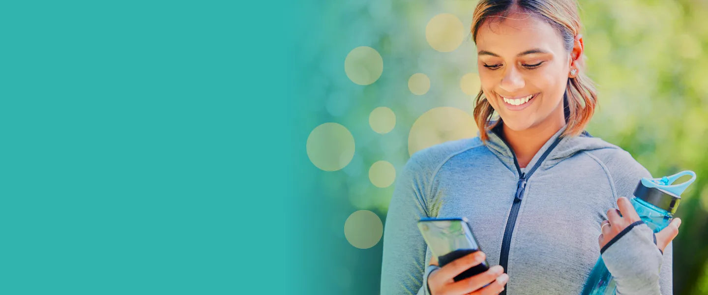 A cheerful young woman in athletic clothing smiling brightly while looking at her smartphone outdoors, holding a reusable water bottle, suggesting a healthy, active lifestyle and highlighting wellness and positivity.
