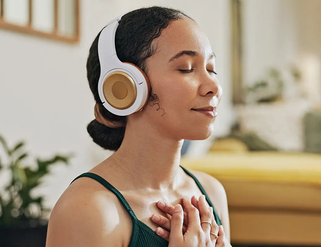 A person wearing white and gold over-ear headphones with their eyes closed, appearing peaceful or meditative. She's wearing a green top and have their hands clasped near their chest.